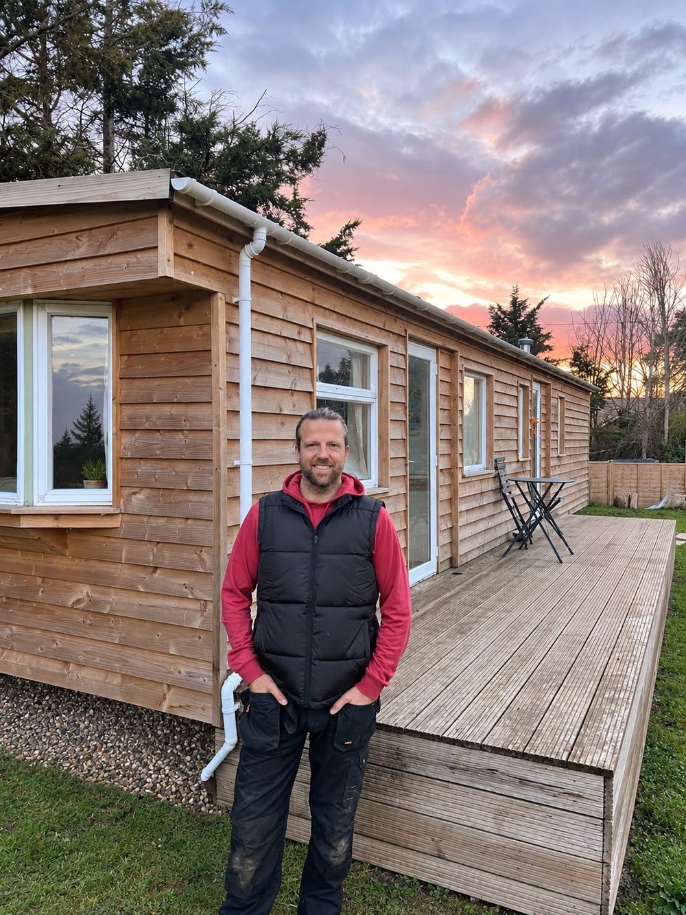 Nick standing beside a timber cabin with a deck at sunset